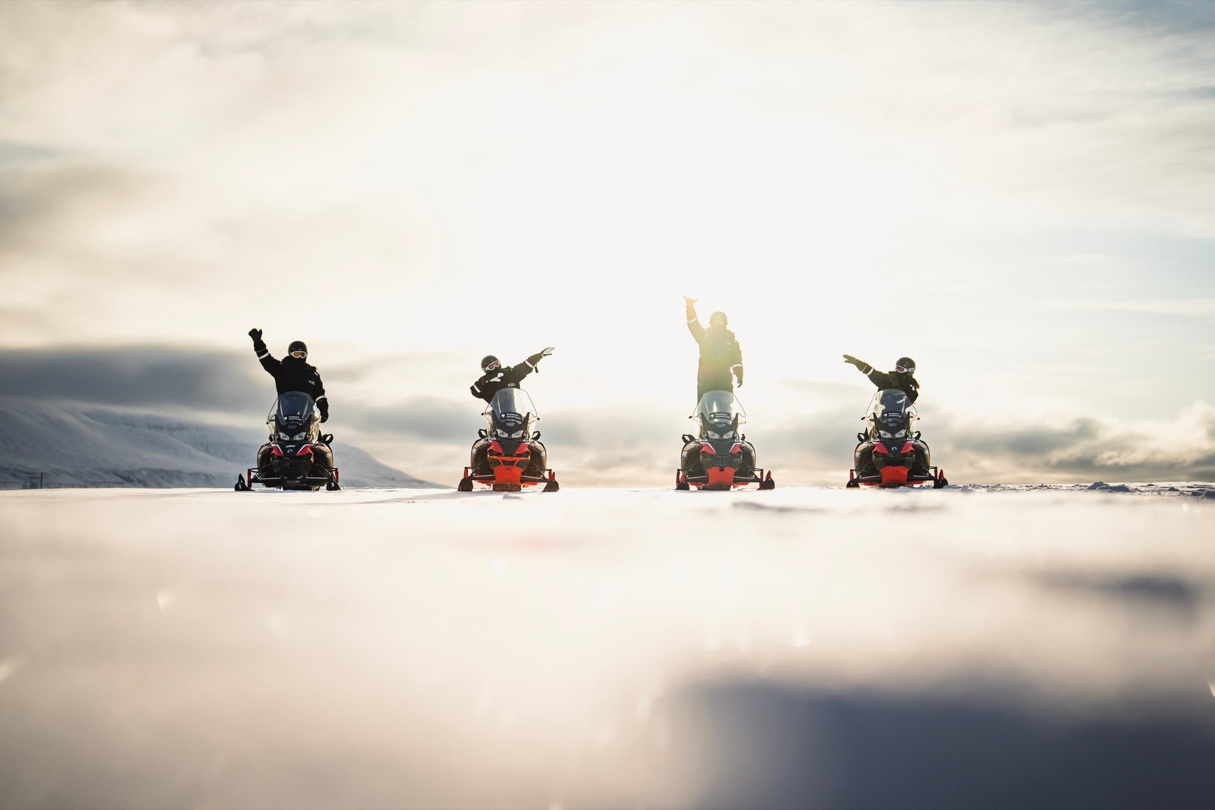 Four snowmobiles halted in a line on a Svalbard snowfield, riders with arms raised into the low arctic sun